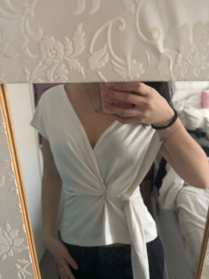 Vit omlottopp med knyt - Snygg vit omlottopp med knyt i midjan och v-ringning. Toppen har korta ärmar och en feminin passform som framhäver midjan. Perfekt att styla med jeans eller kjol för en trendig look.