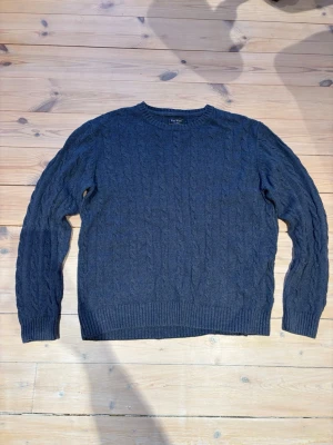 Mörkblå East West tröja - Mysig mörkblå kabelstickad tröja från Oscar Jacobson. Tröjan har rund halsringning, ribbade muddar vid ärmslut och nederkant samt ett klassiskt flätmönster över hela plagget. Perfekt för dig som gillar stilrena och tidlösa plagg.