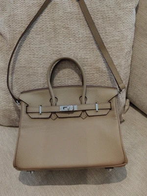 Khaki handväska i skinn med axelrem - Stilren beige handväska i ko skinn med både handtag och avtagbar axelrem. Väskan har silverfärgade metalldetaljer och klassisk låsdetalj framtill. Storlek 25.