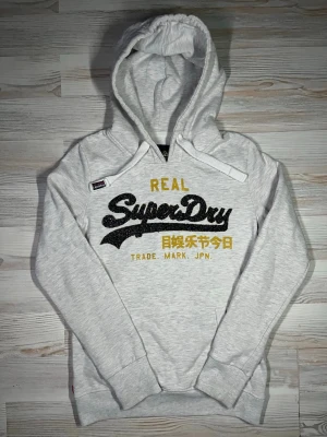 Ljusgrå Superdry hoodie med tryck - Säljer en ljusgrå hoodie från Superdry med stor svart och gul logga samt japansk text på bröstet🤩 Hoodien är i fint skick och passar S🙌🏻 hör av er vid funderingar samt Snabbaffärer