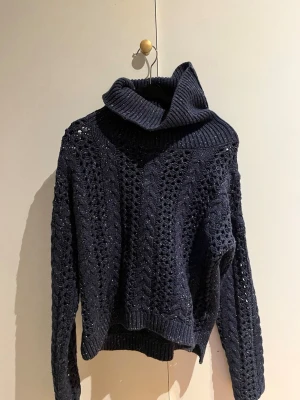 Mörkblå stickad polotröja - Snygg mörkblå stickad tröja med polokrage och hålmönster. Tröjan har lång ärm och ribbade muddar vid ärmslut och nederkant. Perfekt för dig som gillar en avslappnad men trendig stil.