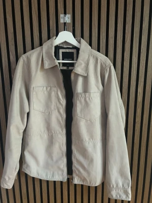 Beige overshirt från Jack & Jones - Stilren beige overshirt från Jack & Jones med två bröstfickor och dragkedja framtill. Overshirten har klassisk krage och är tillverkad i ett mjukt material som ger en avslappnad look. Perfekt att bära över en t-shirt för en trendig stil.