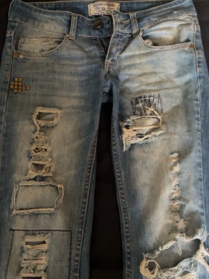 Ljusa slitna jeans från Bershka - Säljer ett par ljusblå jeans från Bershka med kraftiga slitningar och hål på både fram- och baksida. Jeansen har en klassisk femficksmodell, låg midja och detaljer som broderi och patch på vänster ben. Perfekta för en edgy streetstil.