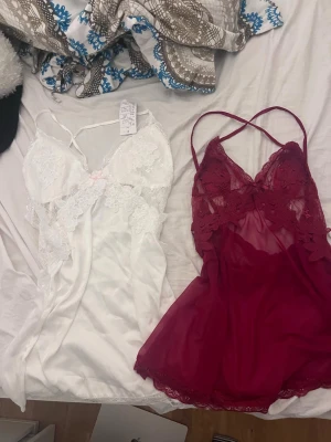 Två spetsnattlinnen i vitt och vinrött - Säljer två nattlinnen i mjuk satin och spets, ett i vitt med blommig spets och liten rosa rosett, och ett i vinrött med transparent mesh och spetsdetaljer. Båda har tunna axelband och romantisk design, perfekta för mysiga kvällar.
