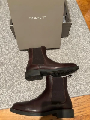 Bruna Chelsea boots från Gant - Snygga mörkbruna Chelsea boots från Gant can i sl grymt stilren design. Skorna är i slätt läder med elastiska sidopaneler och robust sula o har en klassisk siluett. Perfekta för dig som vill ha sl clean och tidlösin look.