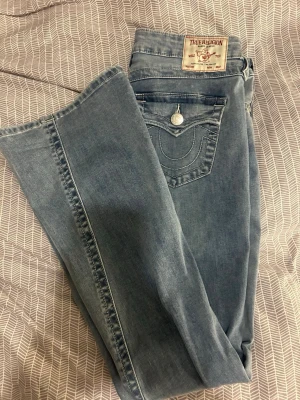 True religion joey - Säljer ett par ljusblå bootcut jeans från True Religion med klassiska kontrastsömmar och ikonisk fickdetalj bak. Jeansen har normal midja och är tillverkade i mjuk denim med lätt tvättad look. Perfekta för en avslappnad och trendig stil.
