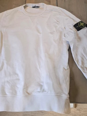 Vit sweatshirt från Stone Island - Säljer en clean vit sweatshirt från Stone Island med klassisk logopatch på vänster ärm. Tröjan har rund halsringning, ribbade muddar och är tillverkad i mjukt bomullsmaterial. Perfekt för en stilren och avslappnad look.
