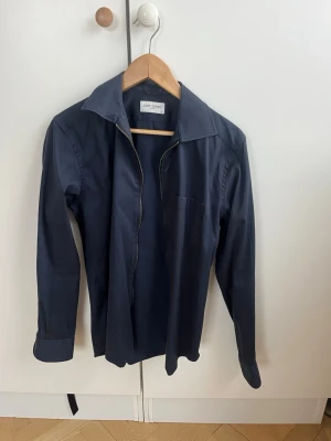 Mörkblå overshirt från John Henric - Snygg mörkblå overshirt från John Henric med lång ärm, klassisk krage och bröstficka. Overshirten  har dragkedja framtill istället för knappar vilket ger en modern touch. Tillverkad i ett mjukt och bekvämt bomullsmaterial. Nypris 1500 kr