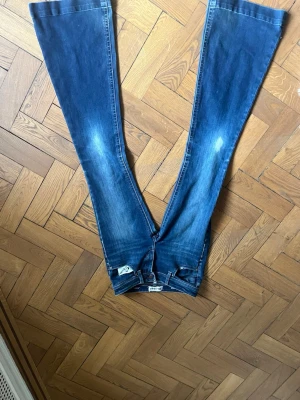 Blå bootcut jeans från Diesel - Säljer ett par blå bootcut jeans från Diesel med klassisk femficksdesign och lätt slitna detaljer på låren. Jeansen har en snygg tvätt och breda ben som ger en retrovibe. Perfekta för dig som gillar en avslappnad men trendig stil. Jätte lowwaist