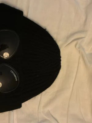 Svart ribbstickad mössa C.P. Company - Svart ribbstickad mössa från C.P. Company med klassiska inbyggda goggles framtill. Mössan är tillverkad i ett mjukt och stretchigt material som sitter skönt på huvudet. Perfekt för dig som vill ha en streetwear-inspirerad look.