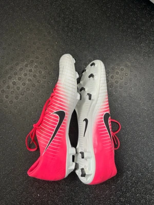 Nike Mercurial rosa/vita fotbollsskor - Säljer ett par Nike Mercurial fotbollsskor i en snygg rosa och vit färgkombination med svarta detaljer. Skorna har snörning, låg profil och fasta dobbar för spel på gräs. Tillverkade i syntetmaterial för lätt känsla och bra bollkontroll.