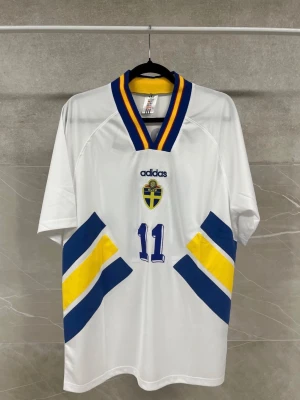 Svensk landslagströja Adidas nr 11 - Vit svensk landslagströja från Adidas med blå och gula detaljer, nummer 11 och namn Brolin på ryggen. Tröjan har en v-ringad krage med blå och gula ränder samt landslagets emblem på bröstet. Perfekt för fotbollsfans som vill visa sitt stöd. Finns i flera storlekar!!! Pris kan diskuteras!!!