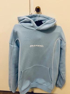Ljusblå hoodie från Dystopia - Ljusblå hoodie från Dystopia med vit logga på bröstet och ryggen. Hoodien har vita kontrastsömmar längs ärmar och kropp, stor känguruficka framtill och en rejäl huva. Tillverkad i mjukt material, perfekt för en chill och stilren look. Passar perfekt för båda S och XS.