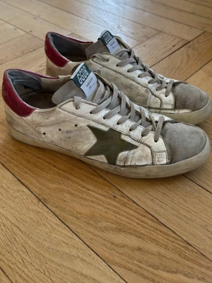 Golden Goose skor - Säljer ett par vita Golden Goose sneakers med olivgrön stjärna på sidan och röd häl sopatch. Skorna har grå snören, slitna sopdetaljer och GG sop target på plösen. Rätt så slitna därav priset. Nypris 5500kr. Inget OG medföljer, pris går att diskutera.