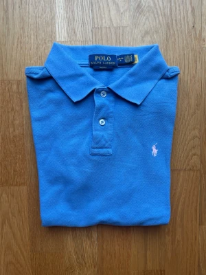 Blå polotopp från Polo Ralph Lauren - Snygg blå piké från Polo Ralph Lauren med klassisk krage och två knappar framtill. Ikonisk broderad logga i rosa på bröstet. Perfekt för dig som gillar stilrena och tidlösa plagg. Storleken är 14-16, passar dig som är 165
