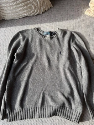 Grå stickad tröja från Polo Ralph Lauren - Klassisk grå stickad tröja från Polo Ralph Lauren med rund halsringning och diskret broderad logga på bröstet. Tröjan har ribbade muddar vid ärmslut och nederkant, perfekt för lager på lager och en clean look.