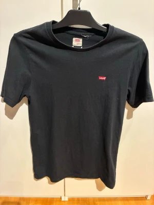 Svart Levi's t-shirt med logga - Klassisk svart t-shirt från Levi's med den ikoniska röda loggan på bröstet. T-shirten har rund halsringning och korta ärmar. Tillverkad i mjuk bomull för en skön och avslappnad stil.