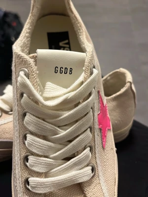 Beige sneakers med rosa stjärna GGDB - Snygga beige sneakers från Golden Goose med en iögonfallande rosa stjärna på sidan. Skorna har vita snören, canvasmaterial och svart sula undertill. Perfekta för dig som vill sticka ut med en cool detalj på fötterna. Saknar låda