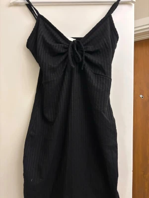 Svart ribbad kortklänning med snörning - Säljer en svart ribbad kortklänning med smala axelband och snygg snörningsdetalj framtill. Klänningen har en figurnära passform och är perfekt för dig som gillar en enkel men trendig stil.