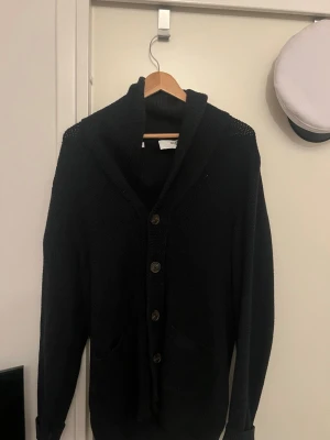 Svart stickad kofta med knappar - Mysig svart stickad kofta från Selected Homme med grovstickad struktur, stora bruna knappar och två fickor framtill. Koftan har en bred sjalkrage och långa ärmar, perfekt för lager på lager under kyliga dagar.