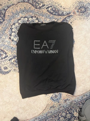 Svart tröja  från Emporio Armani - Snygg svart tröja  från Emporio Armani med stort EA-logotyptryck i vitt på bröstet. Klassisk passform och rund halsringning. Perfekt för dig som gillar stilrena märkeskläder.