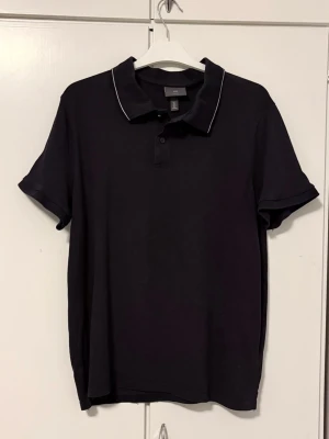 Mörkblå pikétröja med vit rand - Mörkblå pikétröja från H&M med två knappar och en vit rand på kragen.   Köpt begagnad men har själv aldrig använt den.   Storlek L, slim fit