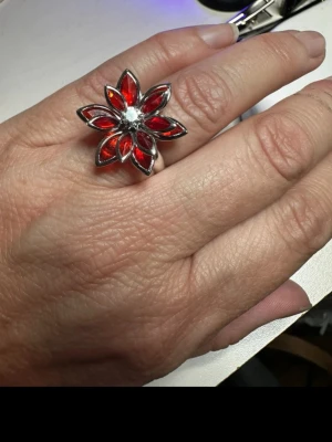 Blommig ring med en röd lotusblomma  - Snygg ring i form av en blomma med sex röda blad och en klar sten i mitten. Ringen har en silverfärgad metallbas och de röda stenarna ger en riktigt cool kontrast. Perfekt för dig som vill sticka ut med ett unikt smycke.