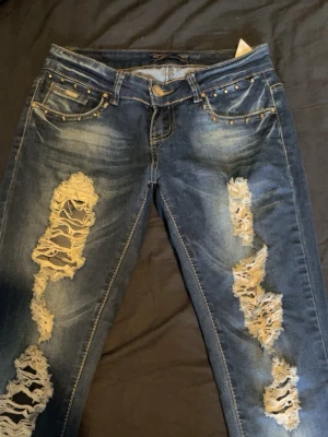 Blå slitna jeans med nitar - Säljer ett par blå jeans med kraftiga slitningar och hål på både fram- och baksida. Jeansen har nitar vid fickorna och en tvättad look som ger en cool, edgy stil. Perfekta för dig som gillar streetwear och vill sticka ut.