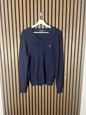 Mörkblå V-ringad tröja Polo Ralph Lauren - Klassisk mörkblå V-ringad tröja från Polo Ralph Lauren i slim fit. Tillverkad i mjuk pima bomull och pryds av den ikoniska broderade loggan i orange på bröstet. Perfekt att bära över en skjorta eller t-shirt för en clean och stilren look.