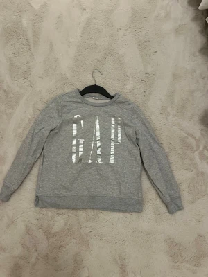 Grå sweatshirt från GAP - Snygg grå sweatshirt från GAP med stor logga i vitt framtill. Tröjan har rund halsringning och långa ärmar. Perfekt för en avslappnad och trendig look. Tillverkad i mjukt material för extra komfort.