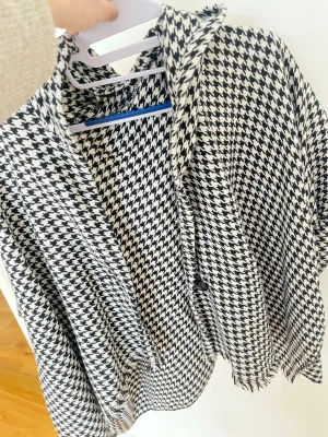 Houndstoothmönstrad sjal i svartvitt - Säljer en stor och mjuk sjal med klassiskt svartvitt hundtandsmönster. Sjalen har fransade kanter och är perfekt att svepa runt sig för en trendig look. Materialet känns som en skön blandning, troligtvis akryl eller ullmix. Enkel att styla till olika outfits.