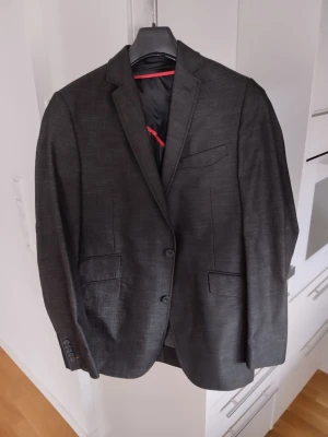 Svart herr kavaj - Stilren svart kavaj från Koppar trans med två knappar framtill, klassisk krage och tre fickor. Insidan har svart foder med röda detaljer. Perfekt för en dressad look och passar till både skjorta och t-shirt.