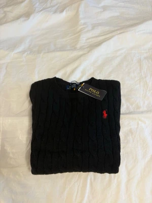 Svart kabelstickad tröja Polo Ralph Lauren - Kabelstickad svart tröja från Polo Ralph Lauren med rund halsringning och röd broderad logga på bröstet. Tröjan har långa ärmar och ribbade muddar vid ärmslut och nederkant. Klassisk och stilren design i mjukt material. Mått:  Längd: 62 cm Axelbredd: 38 cm