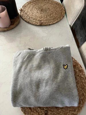 Grå hoodie från Lyle & Scott - Snygg grå hoodie från Lyle & Scott sopassad med klassisk gul logga på bröstet. Hoodien har huva med dragsko och en stor magficka framtill. Tillverkad i mj av stilren sopassad bomullsblandning, perfekt för chill eller streetwear. Stl 12-13 år