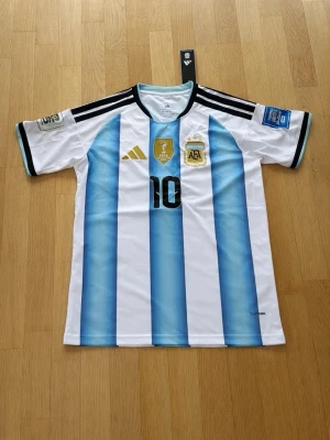 Argentina matchtröja Adidas herr M Messi - Säljer en officiell Argentina matchtröja från Adidas i storlek M. Tröjan har klassiska blå och vita ränder, prod. med ClimaCool-material, svarta axelränder och detaljer i guld. Nummer 10 på bröstet och officiella FIFA- och AFA-emblem. Perfekt för fotbollsfans! Messi
