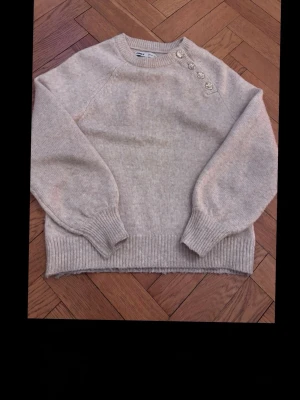 Beige stickad tröja med knappar - Mysig beige stickad tröja med rund halsringning och fyra dekorativa knappar på axeln. Tröjan har ribbade muddar vid ärmslut och nederkant samt ballongärmar för en trendig look. Perfekt för lager på lager och enkel att matcha.