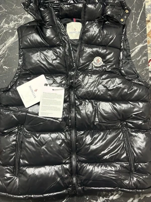 Svart dunväst från Moncler - Snygg svart dunväst från Moncler med glansig finish och ikonisk logga på bröstet. Västen har dragkedja framtill, två stora fickor med dragkedja och hög krage. Insidan har ett serietidningsmönster och etiketter med tvättråd. Perfekt för lager på lager och streetstyle.