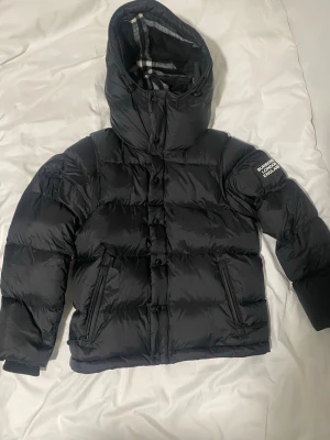 Svart pufferjacka från Burberry - Säljer en svart pufferjacka från Burberry med stor huva och klassiskt rutigt foder. Jackan har dragkedja och knappar framtill, två snedställda fickor med dragkedja och en patch med Burberry-logga på ärmen. Perfekt för kalla vinterdagar.