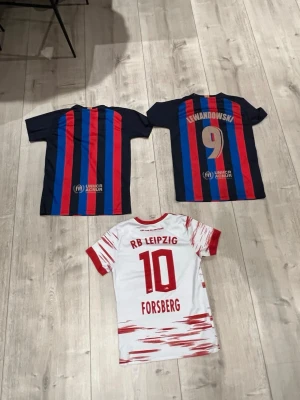 Barcelona och RB Leipzig matchtröjor - Säljer två FC Barcelona matchtröjor med blå och röda vertikala ränder, sponsorloggor och Lewandowski-tryck på ena. En RB Leipzig matchtröja i vitt med röda detaljer, Forsberg-tryck och Red Bull-logga. Perfekta för fotbollsfans som vill representera sina favoritlag.