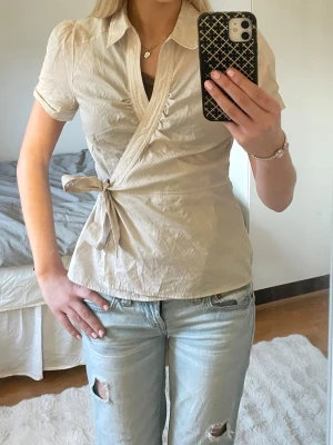 Beige omlottblus med knytband - Snygg beige omlottblus med korta puffärmar och krage. Blusen har ett knytband i sidan och subtila ränder i tyget. Perfekt till jeans ellero kjol för en trendig look. 