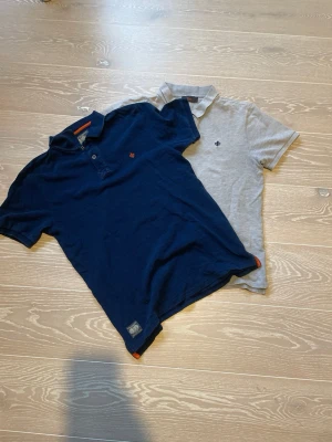 Två pikétröjor i blått och grått - Säljer två stilrena pikétröjor, en marinblå och en ljusgrå. Båda har klassisk krage, knappar framtill och liten broderad logga på bröstet. Perfekta för en avslappnad men snygg look.