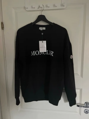 Svart sweatshirt från Moncler - Svart sweatshirt från Moncler med vit logotyp på bröstet och ett broderat Moncler-märke på ärmen/axeln. Klassisk rund halsringning och ribbade muddar. Perfekt för dig som gillar stilrena och exklusiva plagg. Funkar både i storlek S och M