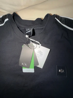  Svart t-shirt Armani Exchange M/L - Svart t-shirt från Armani Exchange med vita detaljer läng volymen på axlarna. Klassisk passform, rund halsringning och diskret A|X-logga på bröstet. Tillverkad i mjuk bomull för en skön känsla. Perfekt relative till jeans eller shorts.