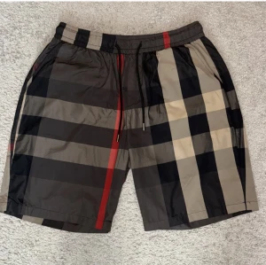 Rutig shorts med snörning från Burberry - Snygga shorts från Burberry med klassiskt rutigt mönster i svart, beige, grått och röda detaljer. De har elastisk midja med snörning och en bakficka. Perfekta för sommaren och ger en clean streetstyle-vibe.