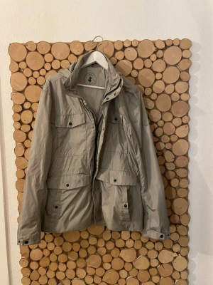 Save the duck field jacket  - Säljer en beige vindjacka från Save The Duck i storlek XL. Jackan är lätt, det sitter en luva på. Men den går att vika in på ett smart sätt som gör jackan riktigt fet. Jackan är en klassisk Field jacket som är riktigt trendigt. Storleken är XL men sitter perfekt på mig som är 190 och väger runt 80 kg. Tvivla inte vid funderingar och priser är inte hugget i sten. Ny pris runt 3000