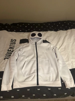 Vit C.P. Company hoodie med glasögon S - Säljer en vit hoodie från C.P. Company med hel dragkedja och ikoniska glasögon i huvan. Hoodien har två stora fickor framtill och svart dragkedja som kontrast. Perfekt för dig som gillar streetwear och unika detaljer passar dig som är 165-169