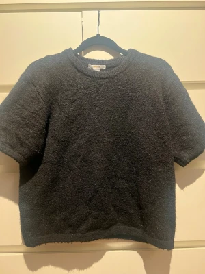 Svart stickad kortärmad topp - Mysig svart stickad topp med korta ärmar och rund hals. Modellen är croppad och har en enkel, stilren design som passar perfekt till jeans eller kjol. Materialet känns mjukt och bekvämt mot huden. Perfekt för alla slags väder.