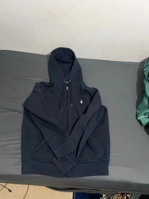 Mörkblå hoodie från Polo Ralph Lauren - Säljer en mörkblå hoodie från Polo Ralph Lauren med dragkedja framtill och två fickor. Klassisk huva med dragsko och den ikoniska vita loggan broderad på bröstet. Perfekt för en chill och stilren look.