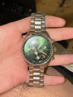 Grön kronografklocka från BOSS - Stilren klocka från BOSS med grön urtavla och kronograf-funktion. Armbandet är i silverfärgad metall med gulddetaljer. Klockan har datumvisning och flera små urtavlor för extra funktioner. Perfekt accessoar för dig som gillar en lyxig och modern look.