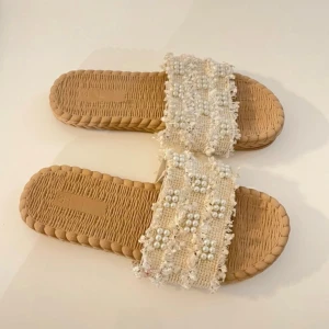 Beige sandaler med pärldetaljer - Snygga beige sandaler med flätad sula och bred rem i tyg dekorerad med vita pärlor och små blommor. Perfekta för en trendig och avslappnad look. Remmen har en fransig kant som ger extra stilpoäng. Passar till sommarens alla outfits.